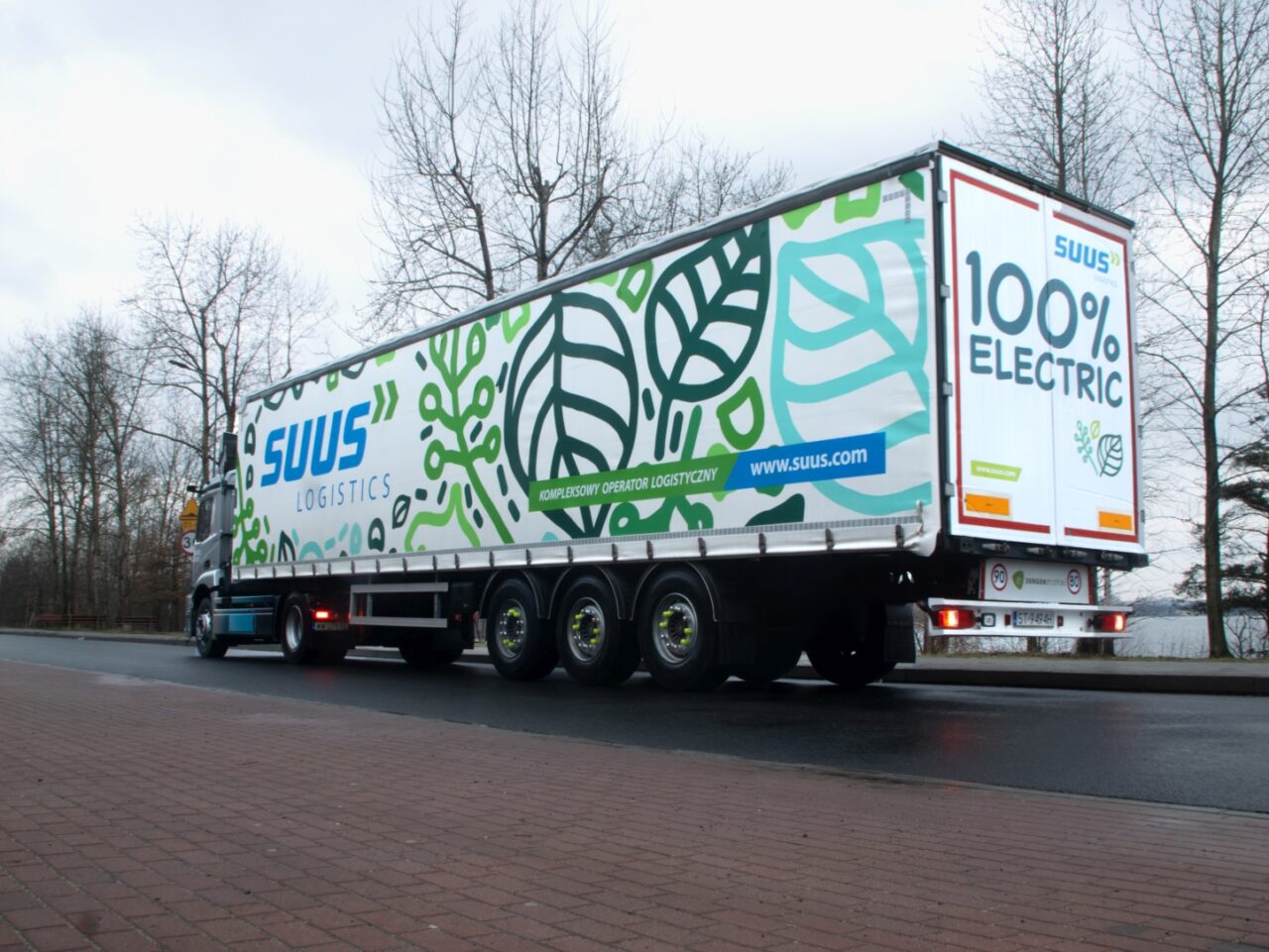 Ciężarówka SUUS Logistics z naczepą w barwach firmowych i napisem "100% ELECTRIC" zaparkowana na poboczu drogi.