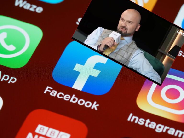 Zbliżenie na ekran smartfona z ikonami aplikacji, na pierwszym planie aplikacja Facebook, a nad nią nachodzący obraz mężczyzny przemawiającego do mikrofonu.