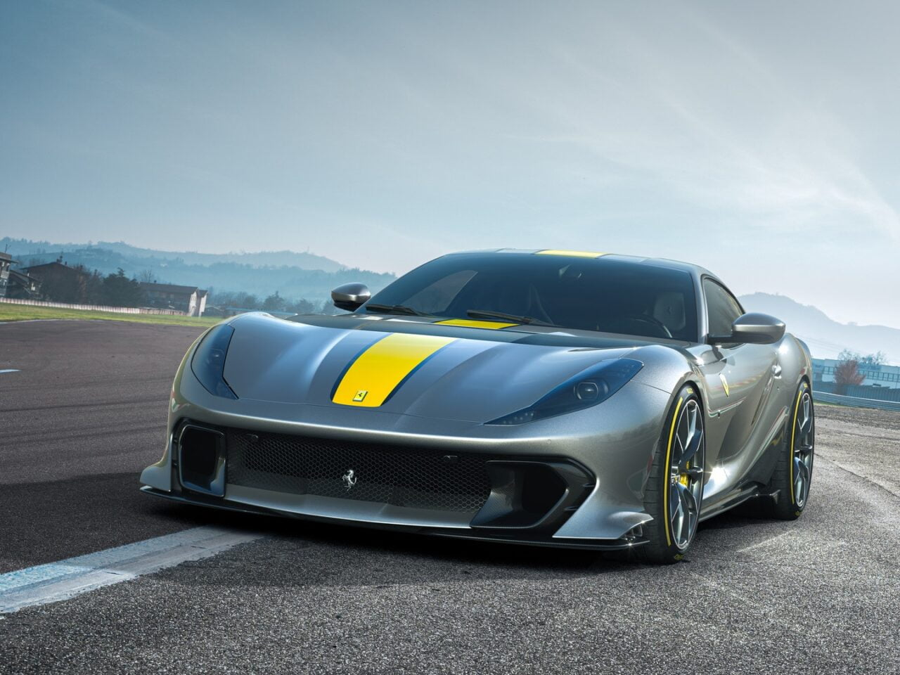 Szary samochód sportowy marki Ferrari z żółtymi akcentami, zaparkowany na asfaltowym torze wyścigowym.