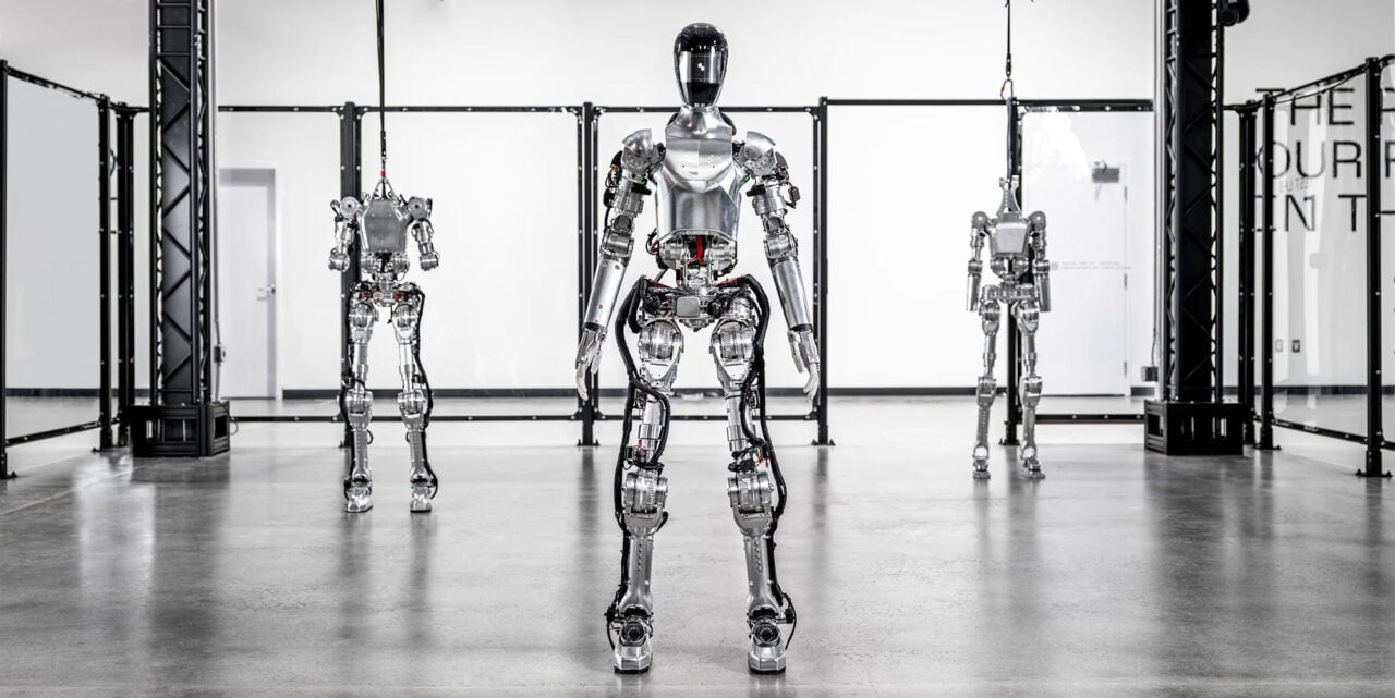 Cztery humanoidalne roboty Figure wystawione w rzędzie w przestronnym wnętrzu laboratorium z błyszczącą podłogą.