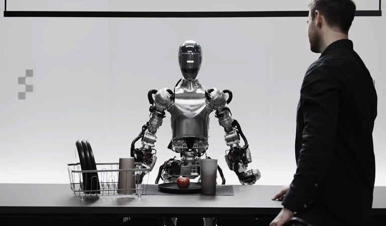 Robot humanoidalny Figure stojący naprzeciwko mężczyzny, wykonujący czynności kuchenne z naczyniami i jabłkiem na blacie.