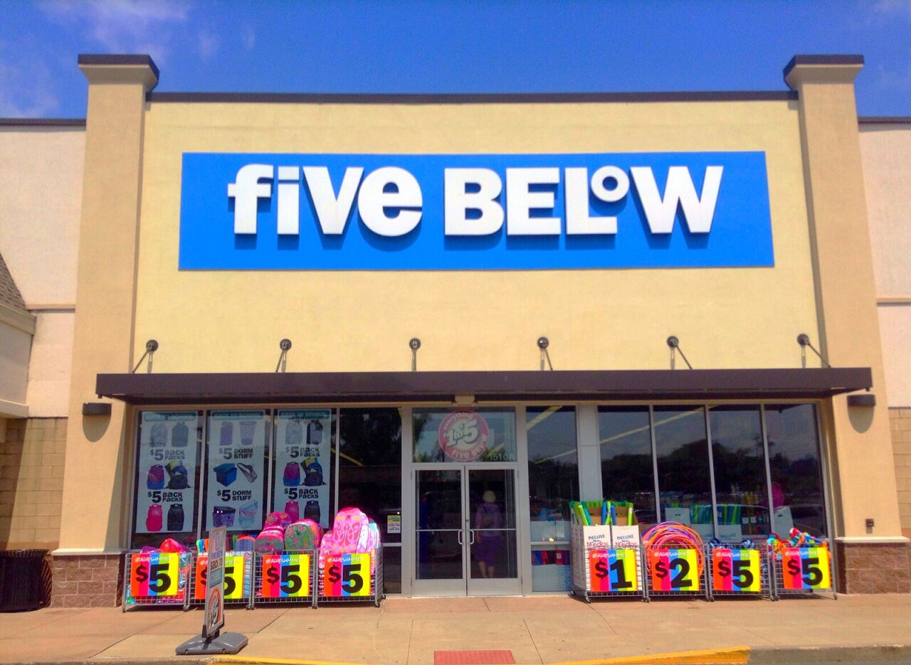 Vista da frente da loja "Cinco abaixo" com um grande logotipo na fachada e vários produtos coloridos na frente da entrada com etiquetas de preços bem visíveis, incluindo "5$", "1$" e "2$".