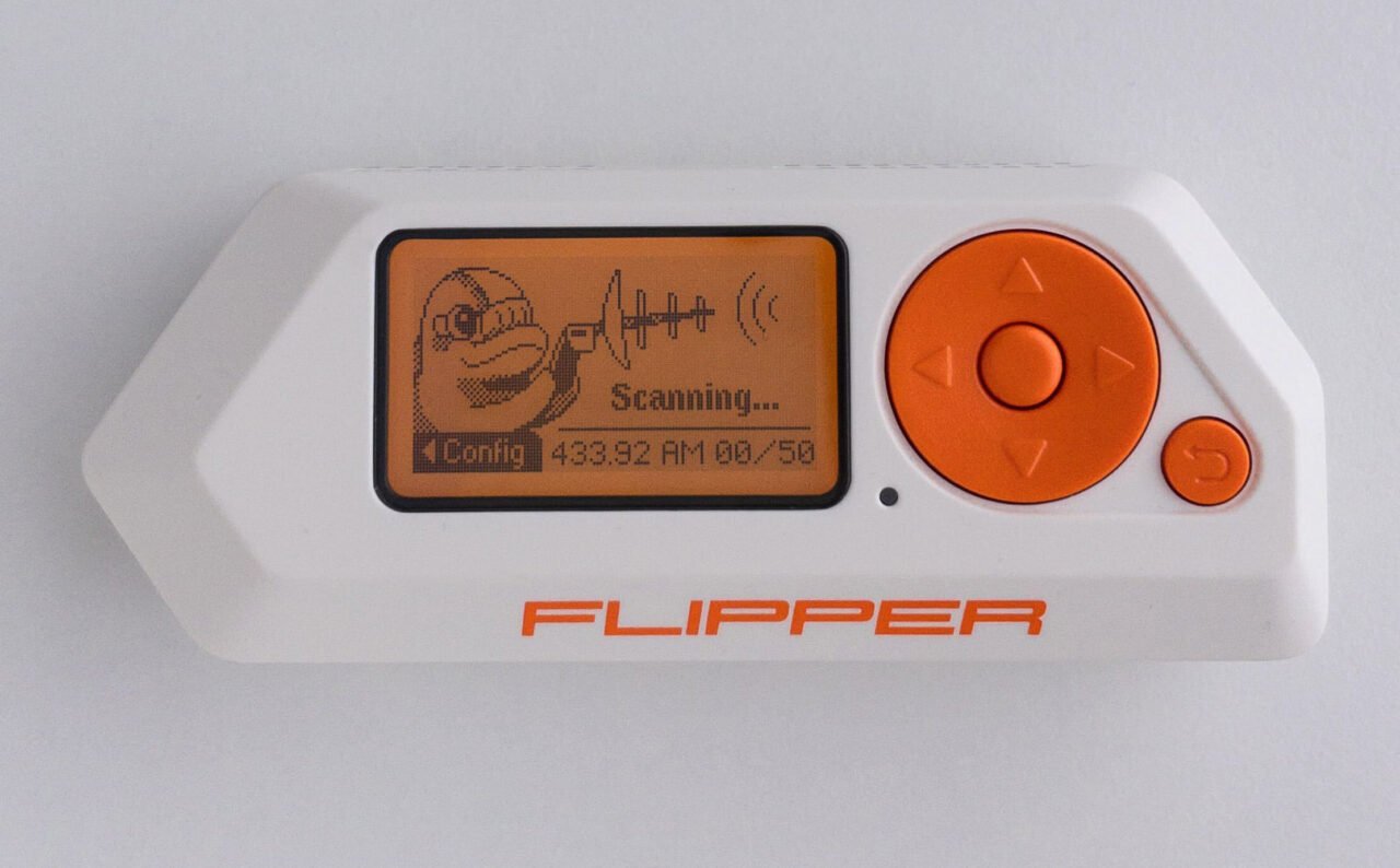 Urządzenie Flipper Zero z wyświetlaczem LCD, na którym widać grafikę uśmiechniętej twarzy i napis "Scanning...", oraz z przyciskami nawigacyjnymi w kolorze pomarańczowym.