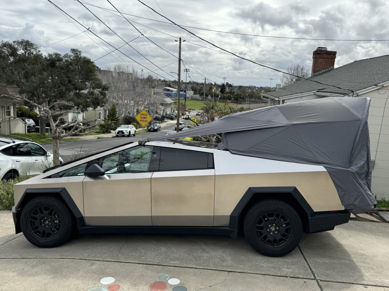 Namiot Tesla Basecamp. Samochód o wyraźnym, kanciastym kształcie z osłoną na tylnej części zaparkowany przy ulicy na przedmieściach; tło z domami i roślinnością.