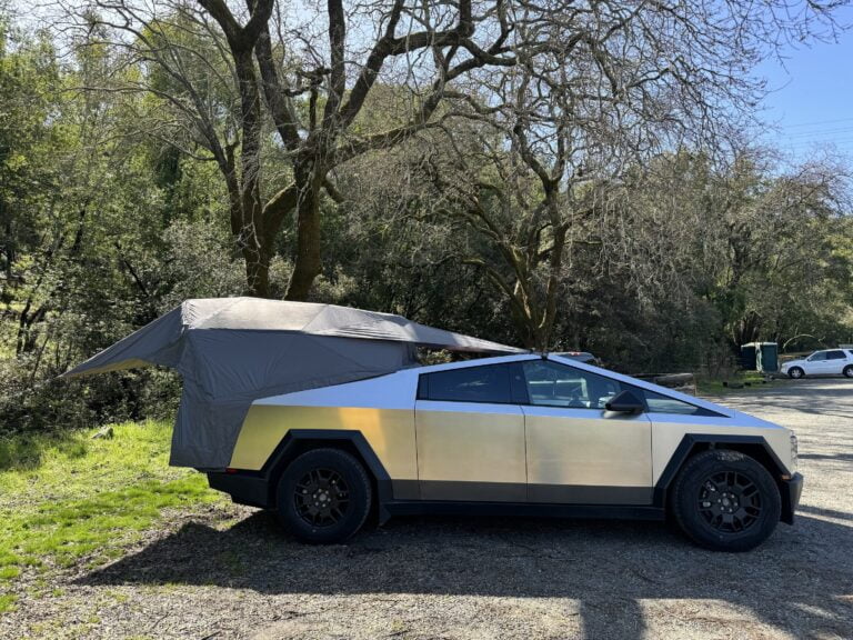 Samochód o futurystycznym wyglądzie z przyczepionym namiotem na zewnątrz, zaparkowany w leśnym terenie.