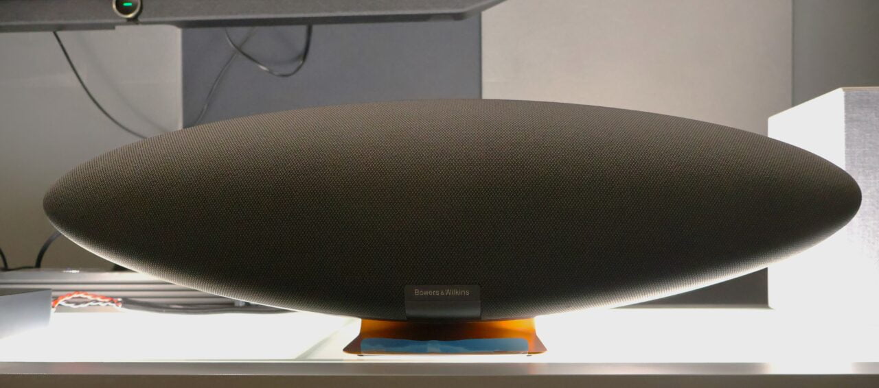 Czarny, owalny głośnik marki Bowers & Wilkins umieszczony na półce przed telewizorem.