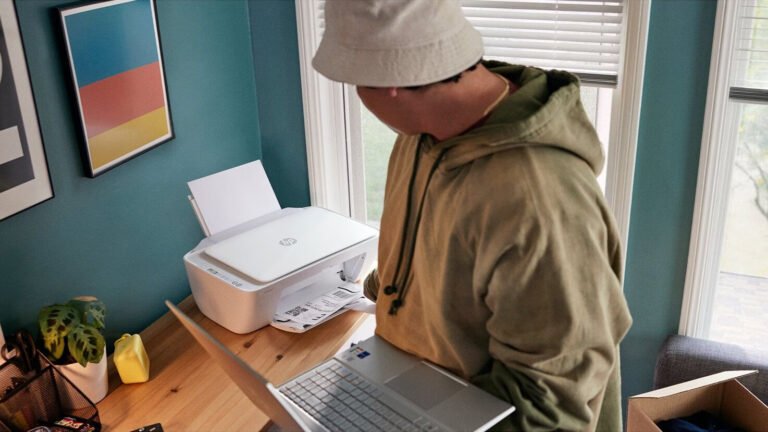 Mężczyzna w beżowej bluzie z kapturem używa drukarki obok laptopa w domowym biurze.