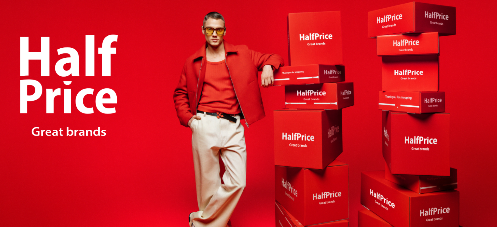 Mężczyzna w czerwonym garniturze pozuje obok wieży z czerwonych pudeł z napisem "HalfPrice Great brands", na czerwonym tle z tekstem "HalfPrice Great brands".