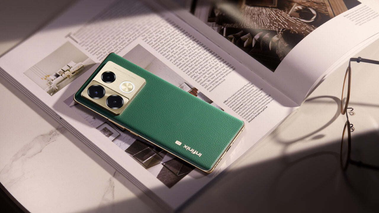 Smartfon w zielonym etui leży na otwartej książce obok okularów na marmurowym blacie.