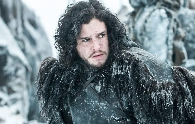 Kit Harington jako Jon Snow w serialu Gra o Tron. Mężczyzna z długimi, czarnymi włosami i brodą, ubrany w ciężki, pokryty śniegiem futrzany płaszcz, stoi na śnieżnym tle, prezentując poważny wyraz twarzy.