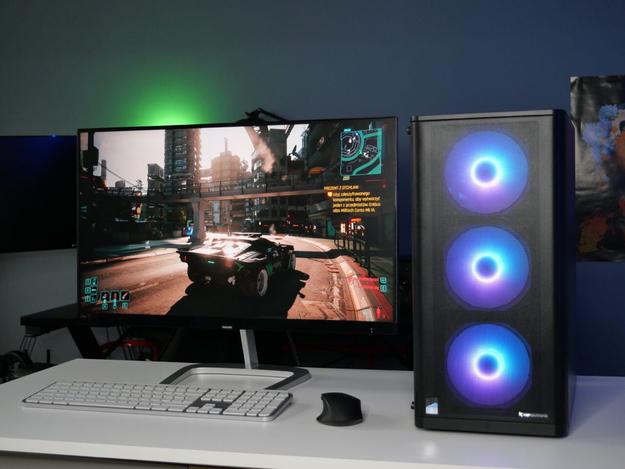 Zestaw komputerowy z podświetlaną obudową PC i dwoma monitorami, na których wyświetlana jest gra wideo, na biurku z białą klawiaturą i czarną myszką. W tle oświetlenie RGB.