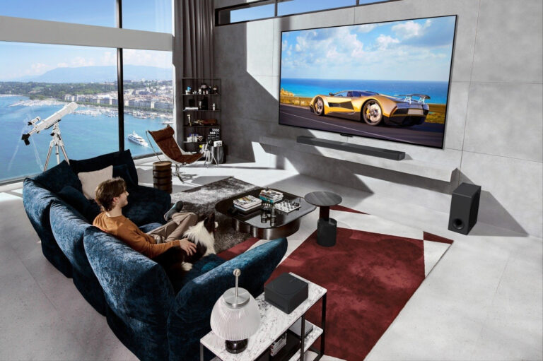 Osoba siedząca na granatowej kanapie w nowoczesnym salonie z dużymi oknami, przez które widać widok na morze i przybrzeżne miasto, ogląda na dużym telewizorze LG OLED film o żółtym samochodzie sportowym; w pomieszczeniu znajduje się teleskop, stół z książkami i dywan.