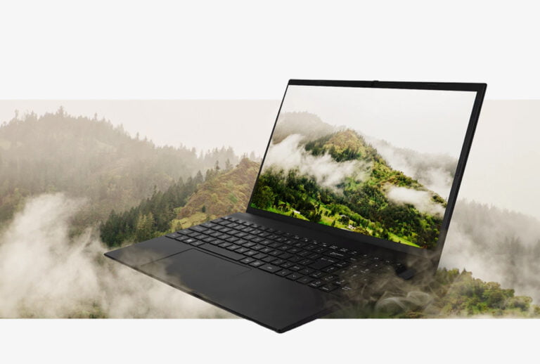 Laptop z wyświetlaczem, na którym kontynuuje się obraz mglistego, zielonego krajobrazu górskiego, tworząc efekt przejścia między rzeczywistym urządzeniem a zaaranżowanym tłem.
