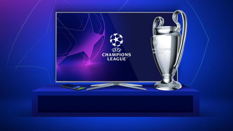 Telewizor wyświetlający logo Ligi Mistrzów UEFA, obok którego znajduje się model pucharu, smartfon i piloty na niebieskim stoliku.