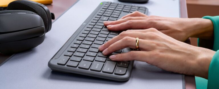 Ręce osoby piszącej na bezprzewodowej klawiaturze, obok słuchawki na biurku.