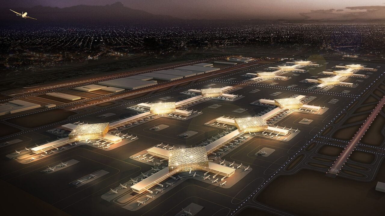 Lotnisko nocą widziane z lotu ptaka, oświetlone terminale i pasy startowe, z samolotem w powietrzu i rozbłyskującymi światłami miasta w tle.