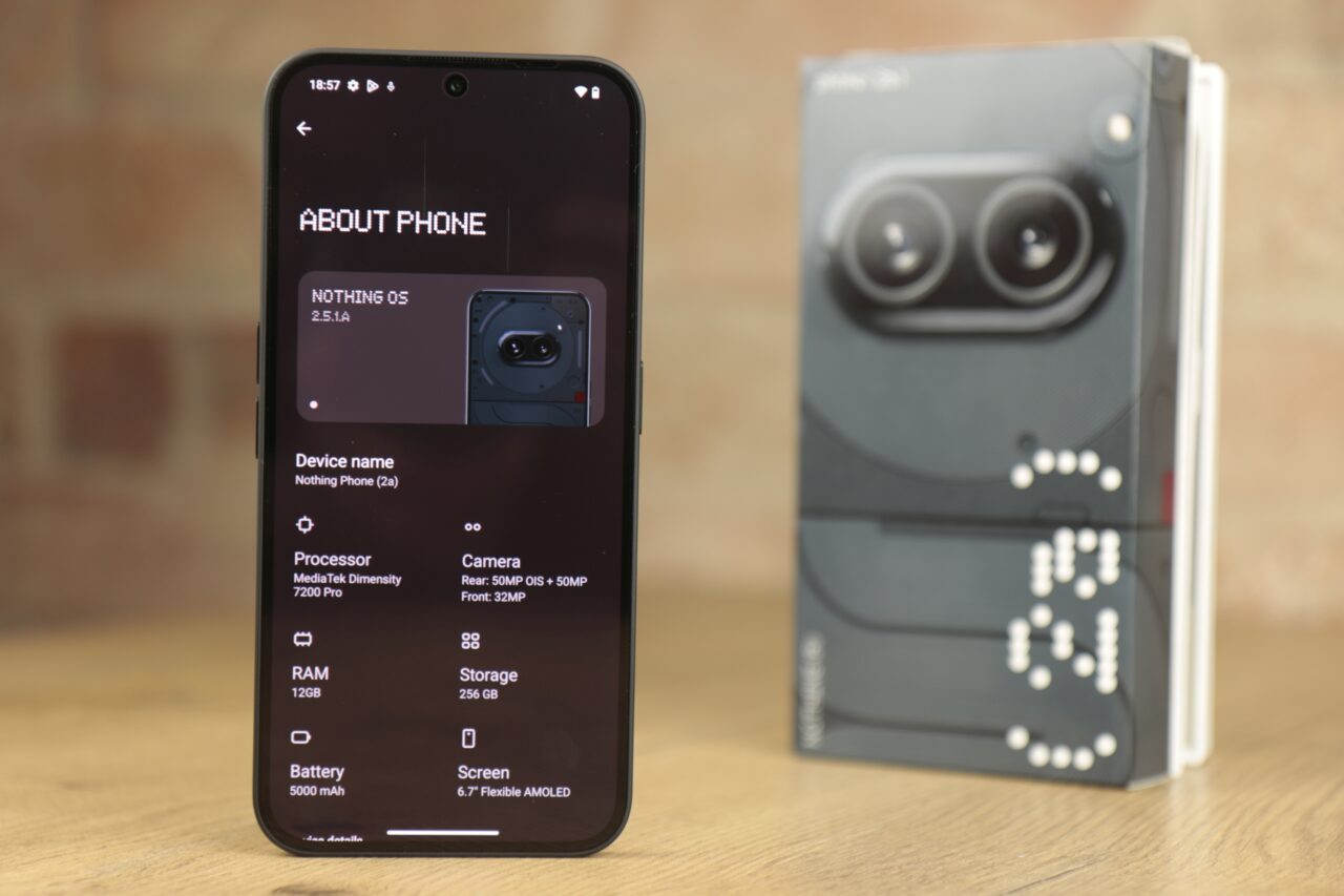 Czarny smartfon Nothing Phone 2a wyświetlający ekran z informacjami o telefonie na pierwszym planie, a w tle pudełko produktu z niewyraźnym obrazem.