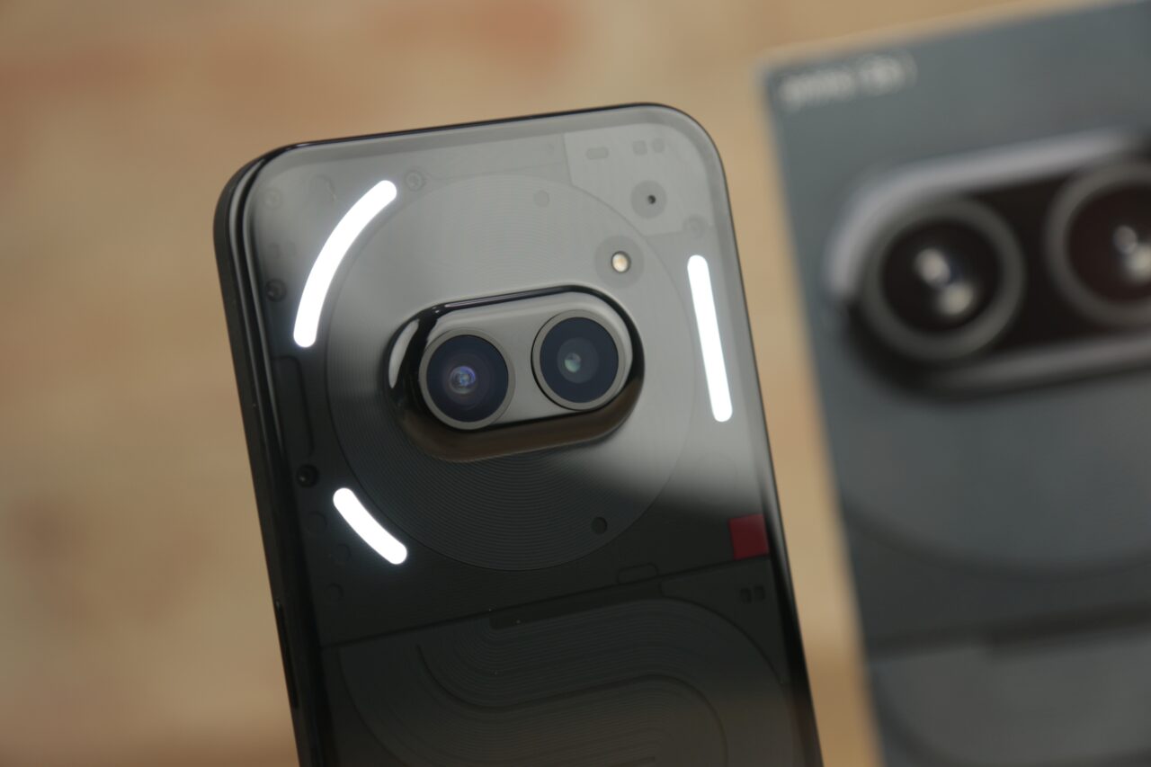 Aparat fotograficzny smartfona Nothing Phone (2a) z podwójnym obiektywem i pierścieniem światła LED, pierwszy plan częściowo rozmyty, w tle niewyraźnie opakowanie produktu.
