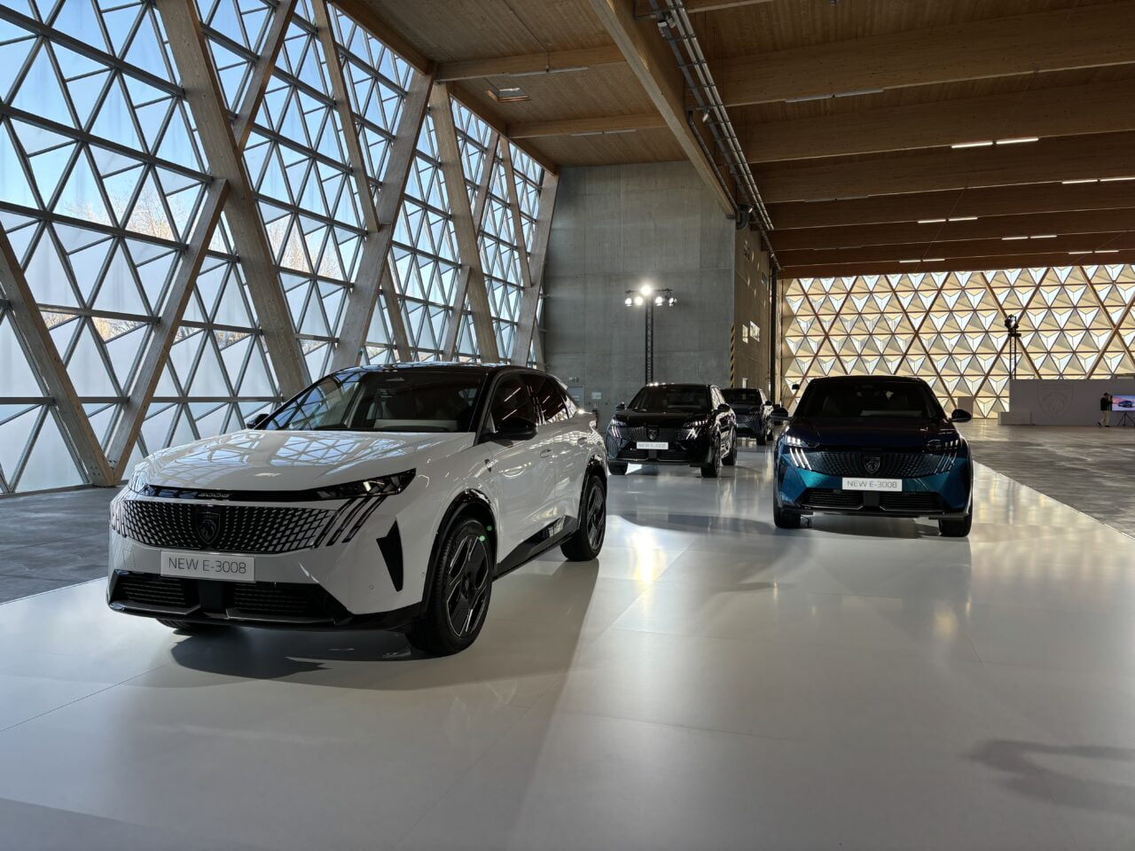 Pokaz salonu samochodowego z kilkoma nowymi modelami samochodów, w tym przedni widok białego samochodu oznaczonego jako NEW E-3008. Wnętrze salonu jest jasne, z dużymi, geometrycznymi oknami i drewnianym sufitem.