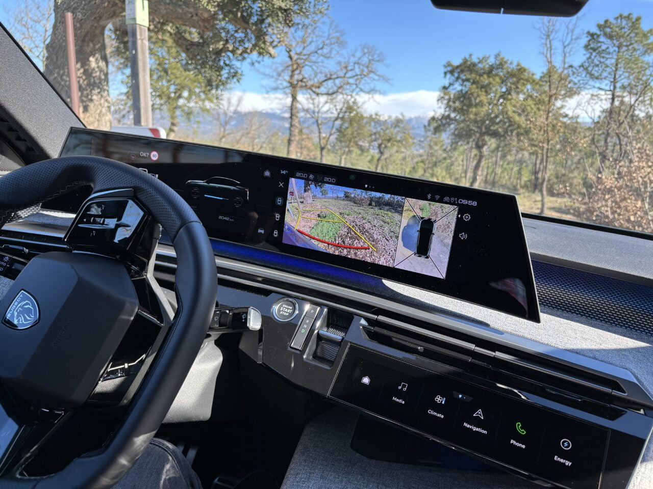 Wnętrze samochodu z widokiem na wielofunkcyjną kierownicę i szeroki ekran dotykowy systemu nawigacyjnego wyświetlający obrazy z kamer zewnętrznych, w tle widoczny krajobraz z drzewami.