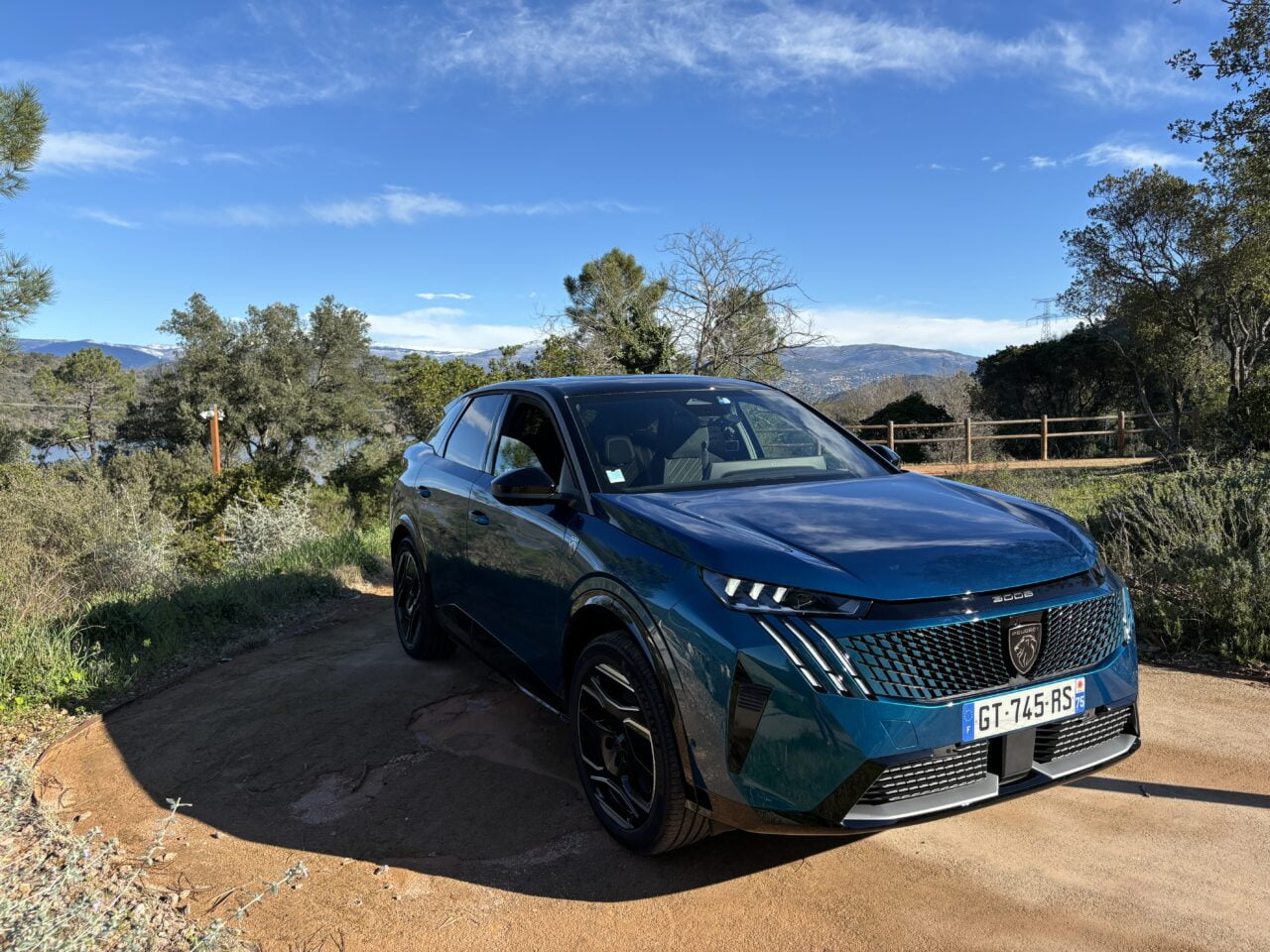 Niebieski samochód marki Peugeot zaparkowany na żwirowej drodze z widokiem na górzysty krajobraz i drzewa w słoneczny dzień.