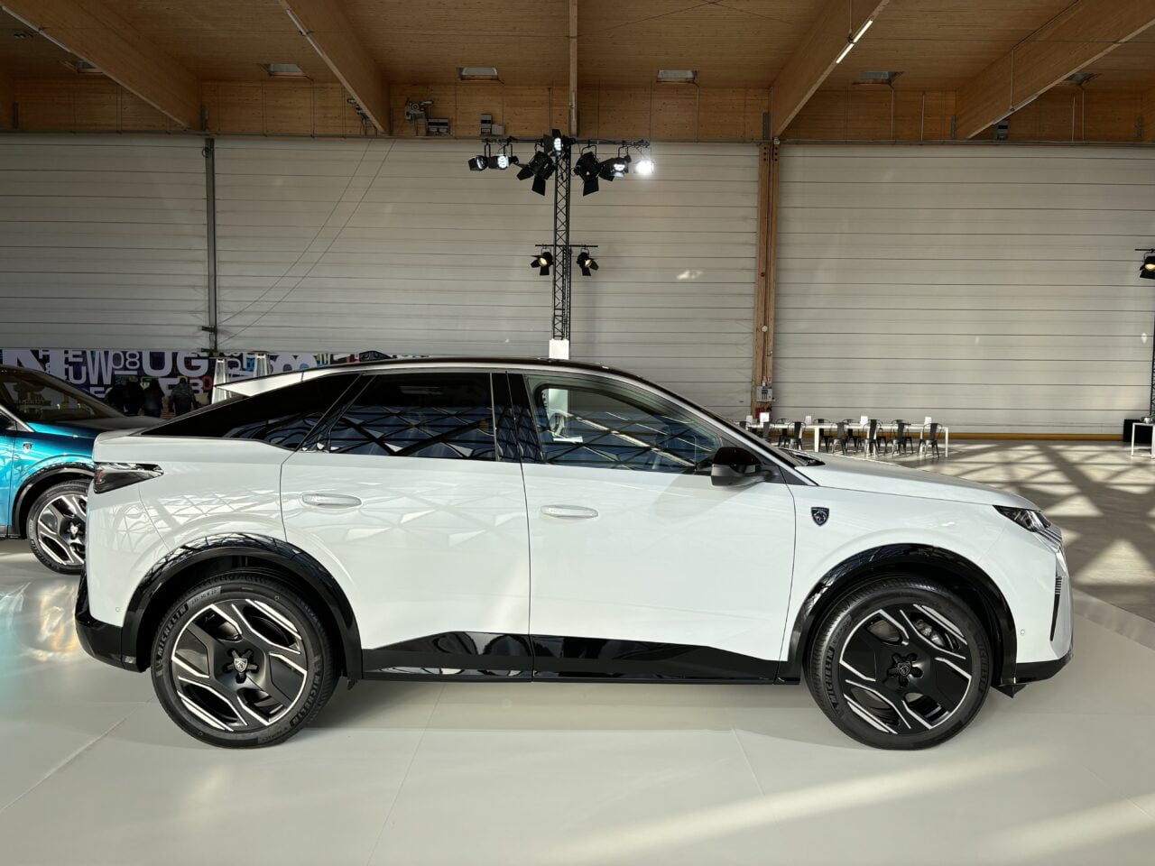 Biały samochód elektryczny SUV z charakterystycznym wzorem na bocznych drzwiach, wystawiony w jasnym salonie z drewnianym stropem.