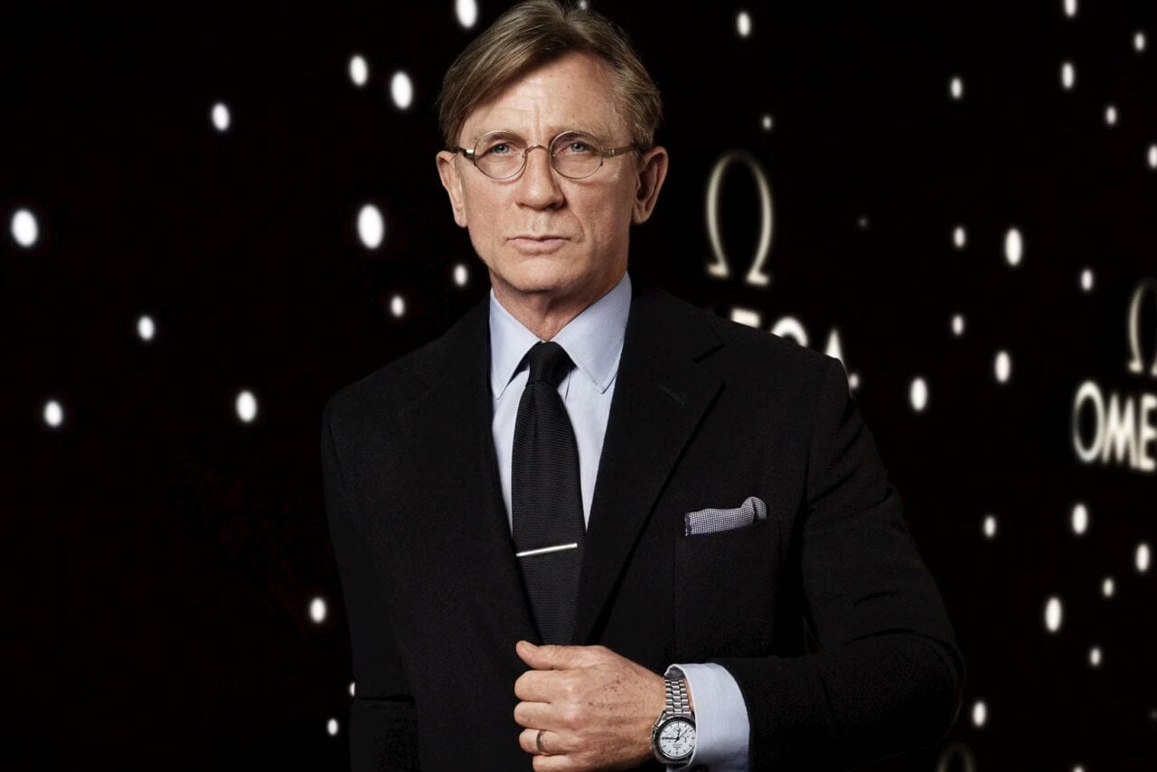 Daniel Craig w eleganckim, ciemnym garniturze z krawatem i okularami, stojący na tle z rozmytymi światłami. Na nadgarstku nosi srebrny zegarek.