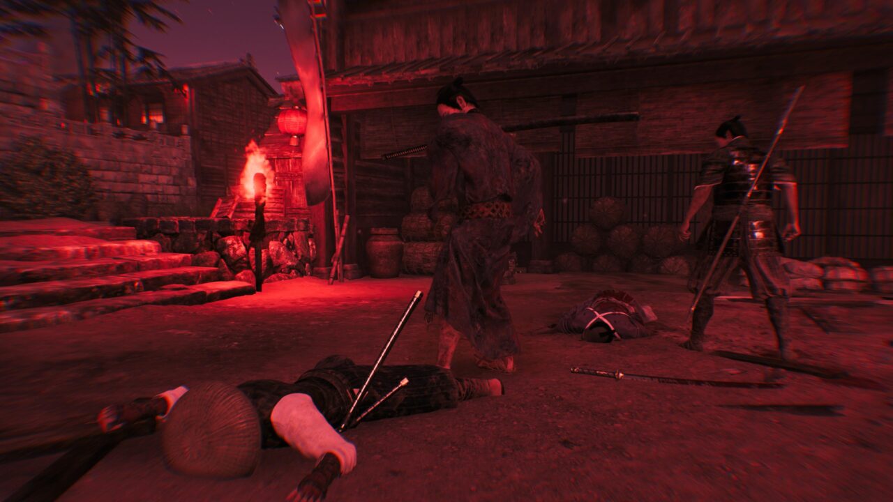 Scena nocna w grze komputerowej przedstawiająca dwóch wojowników w przemierzając wioskę, z przewróconym ciałem i bronią w tle, przy czerwonym oświetleniu pochodni.