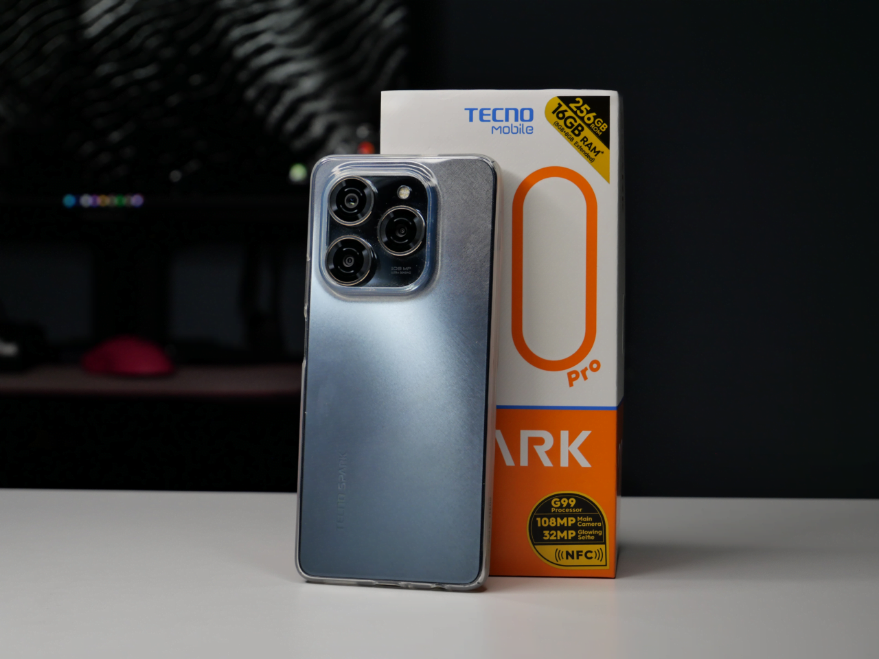 Smartfon TECNO SPARK 8 Pro oparty na tyle obudowy, z zestawem potrójnych aparatów, obok pudełka produktowego z napisami o charakterystyce technicznej, w tle rozmazane biurko z ciemnymi odcieniami.
