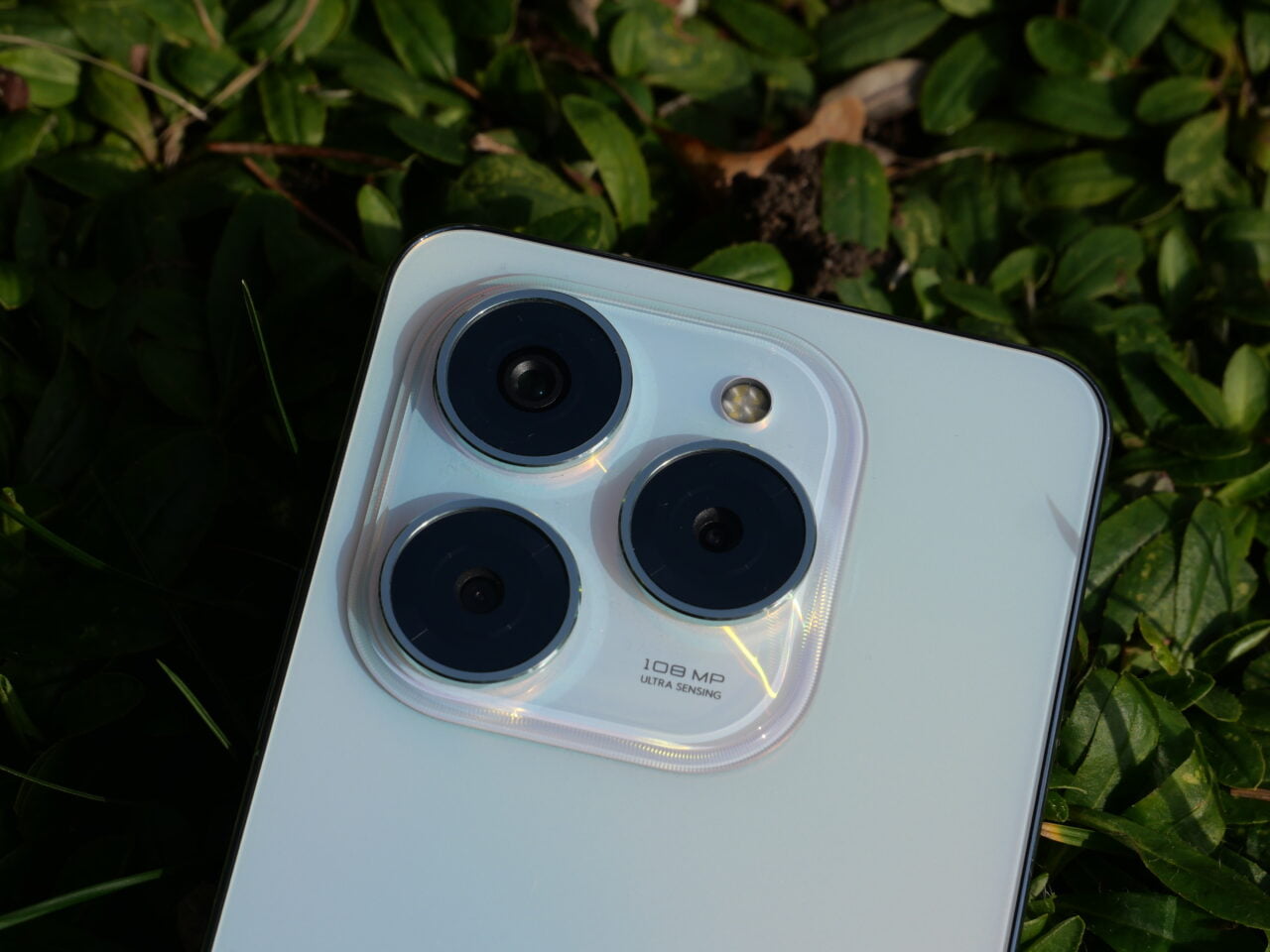 Smartfon leżący na liściach, ukazujący szczegół aparatu z trzema obiektywami i etykietą "108 MP ULTRA SENSING".