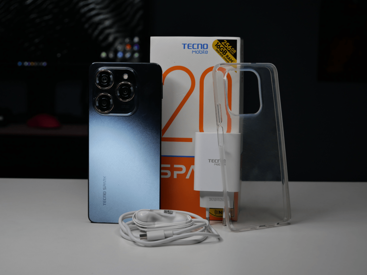 Smartfon Tecno Spark z niebieskim gradientem na plecach i poczwórnym aparatem, pudełko produktu, przezroczyste etui na telefon, ładowarka i biały przewód USB.