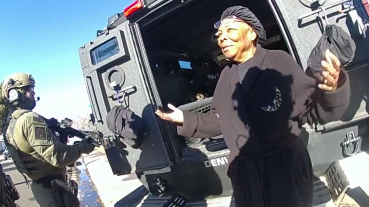 Starsza kobieta w czarnej bluzie i chustce na głowie stoi obok otwartych drzwi czarnego pojazdu policyjnego, rozmawiając z zamaskowanym funkcjonariuszem w zielonym mundurze taktycznym i hełmie, który stoi przed nią wyposażony w broń.