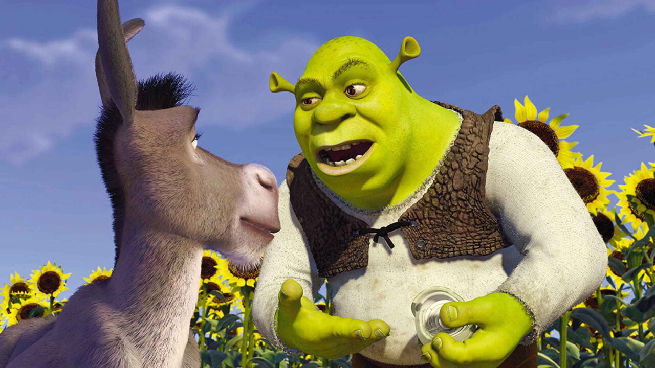 Postać animowana z filmu Shrek przypominająca ogra rozmawia z osłem, w tle pole słoneczników i niebieskie niebo z białymi chmurami.