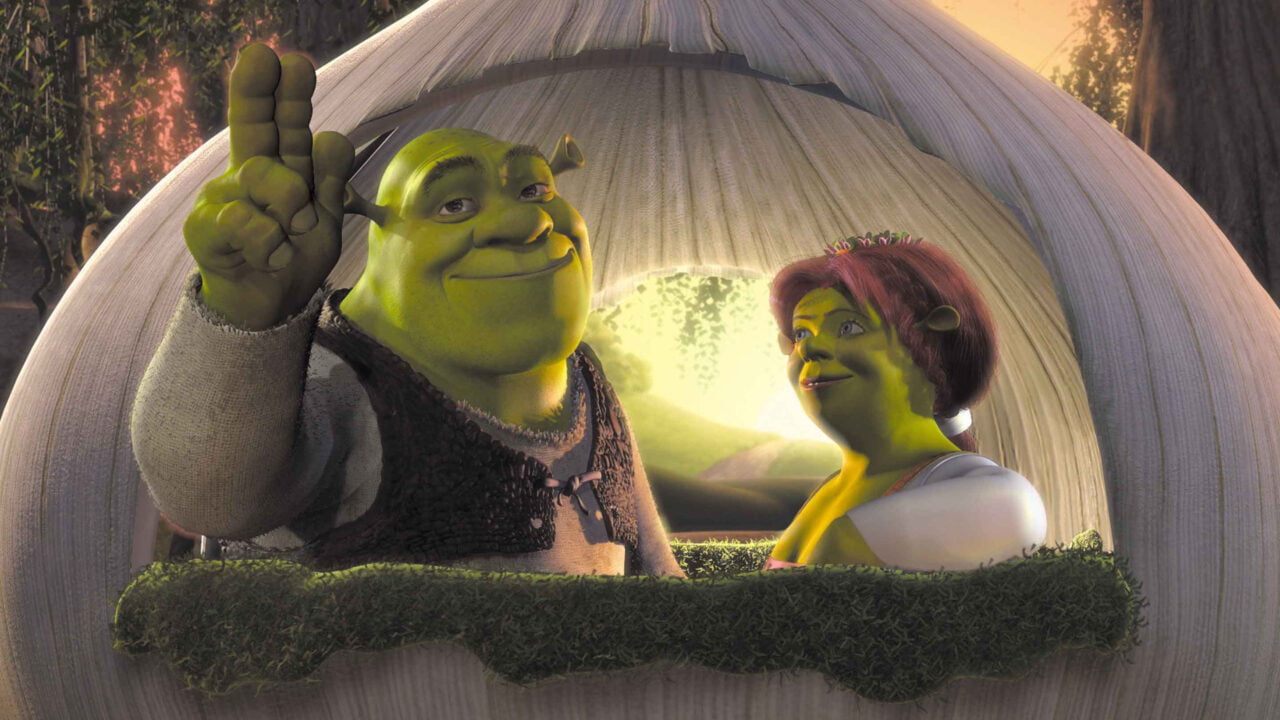 Zdjęcie do artykułu o filmie Shrek 5. Shrek i Fiona stoją pod drzwiem w kształcie łuku, Shrek macha ręką w geście powitania, oboje uśmiechają się.