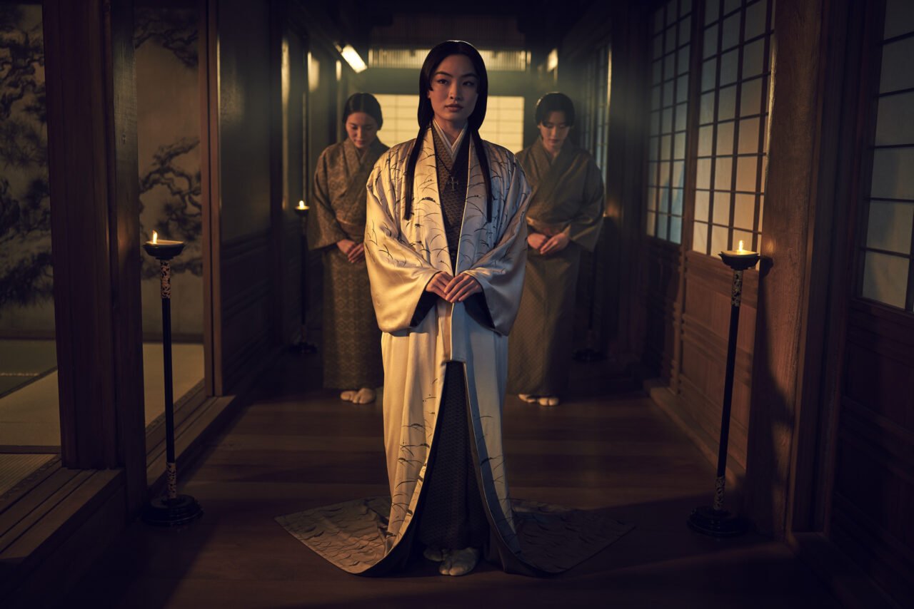 Cztery osoby w tradycyjnych japońskich strojach stoją wzdłuż drewnianego korytarza w tradycyjnym japońskim domu z drzeworytami na ścianach i świecami jako oświetlenie. Osoba na pierwszym planie nosi jasny kimono i stoi z złożonymi rękami, patrząc uważnie w kamerę.