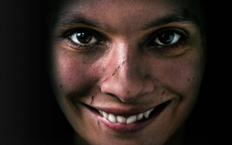 Portret osoby z uśmiechniętą twarzą, widoczne są jedynie oczy, nos i usta, reszta twarzy zanurzona w cieniu.