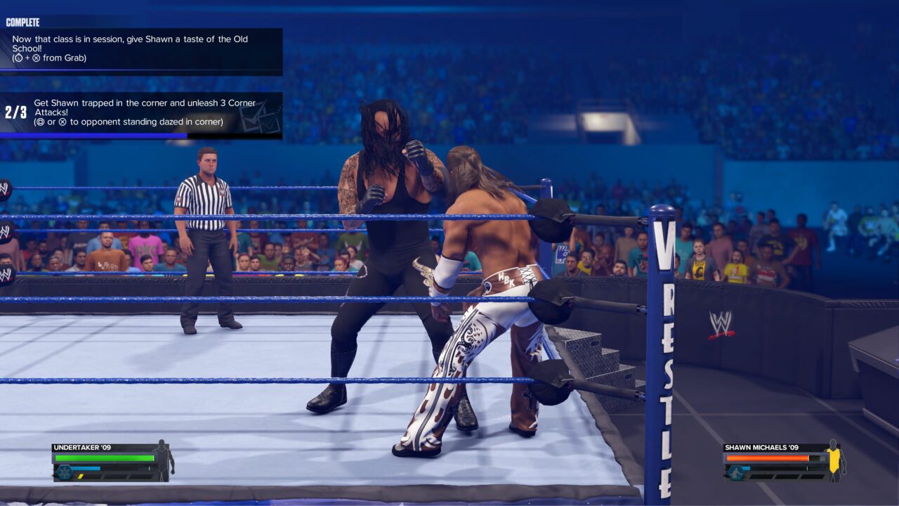 Gra wideo WWE 2K24 z Undertakerem '09 atakującym Shawna Michaelsa '09 w ringu podczas walki, z widownią w tle i elementami interfejsu użytkownika wskazującymi instrukcje i stan zdrowia postaci.