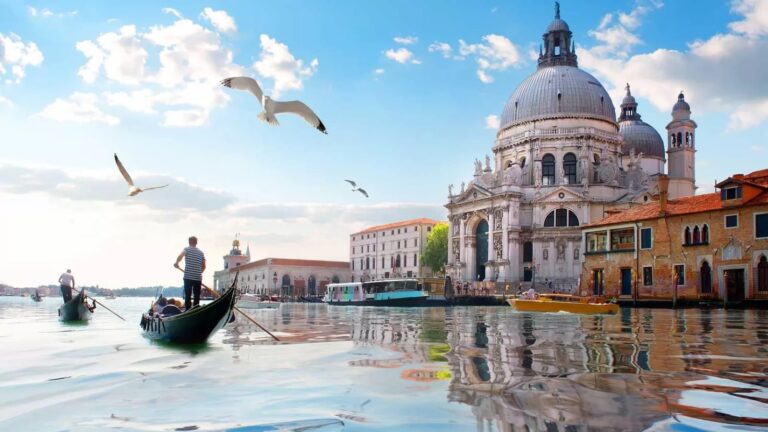 Gondole na spokojnych wodach Kanału Grande w Wenecji, z widokiem na Bazylikę Santa Maria della Salute w słoneczny dzień, z mewami latającymi na niebieskim niebie.