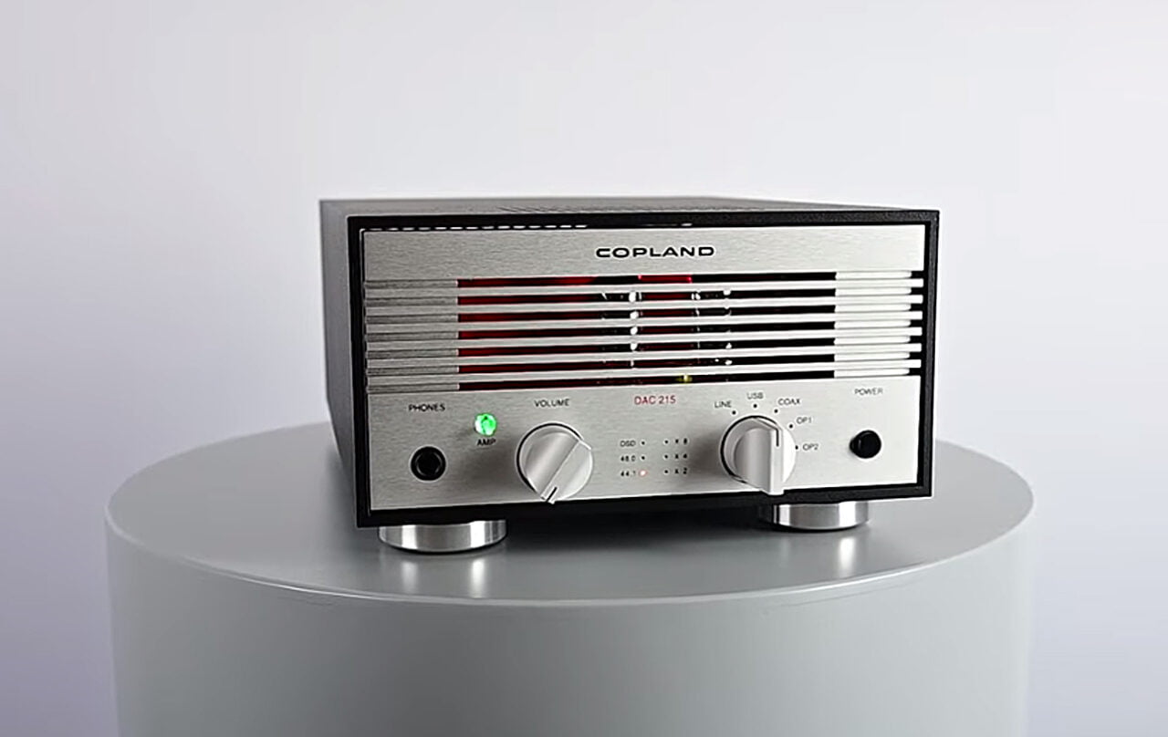 Wzmacniacz audio marki Copland umieszczony na białym stoliku, z aluminiowym frontem, pokrętłami do regulacji głośności i wybierania źródła dźwięku oraz wskaźnikami LED.