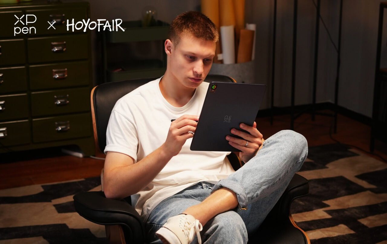 Tablet XPPen. Młody mężczyzna siedzący w fotelu i trzymający cyfrowy tablet z pisakiem, w pomieszczeniu z zielonym kredensem i geometrycznym dywanem.