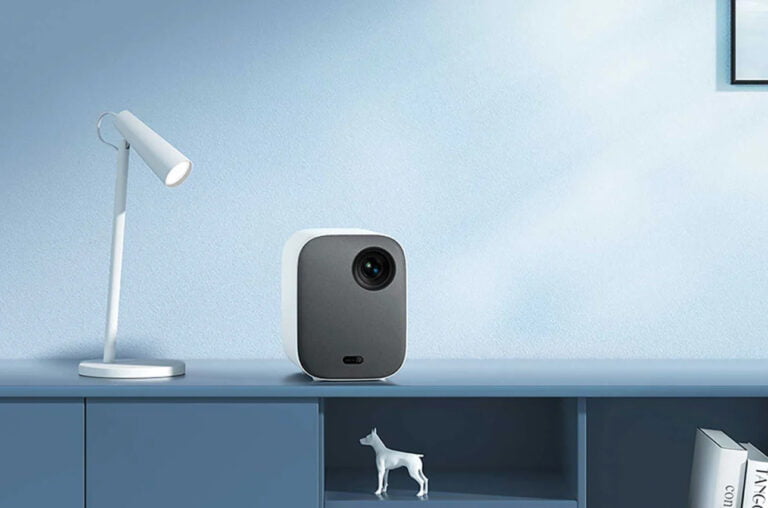 Stylowy, nowoczesny biurkowy gabinet z białą lampką biurkową i projektorem Xiaomi na niebieskiej komodzie z małą figurką psa na półce i obrazem na ścianie.