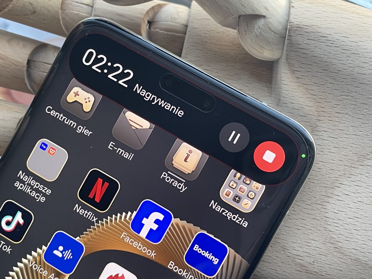 Część przedniego panelu czarnego smartfona z włączonym ekranem pokazującym aplikacje, w tle ręka manekina drewnianego. Ekran wyświetla godzinę 02:22, różne ikony aplikacji i czerwony przycisk nagrywania na górnym pasku.