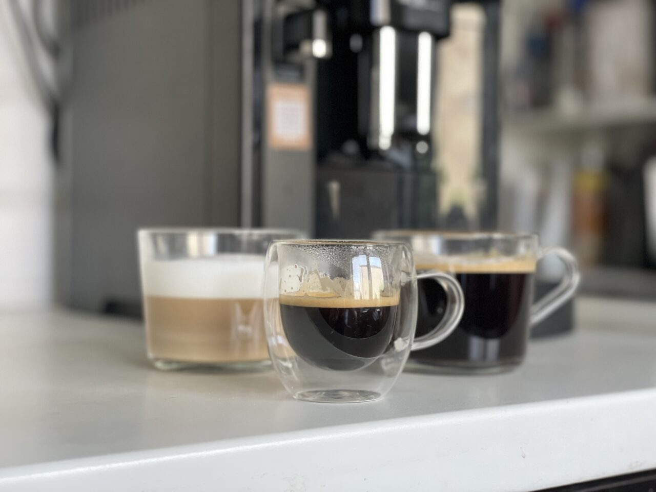 Trzy filiżanki z kawą o różnych konsystencjach na blacie kuchennym z ekspres do kawy w tle.