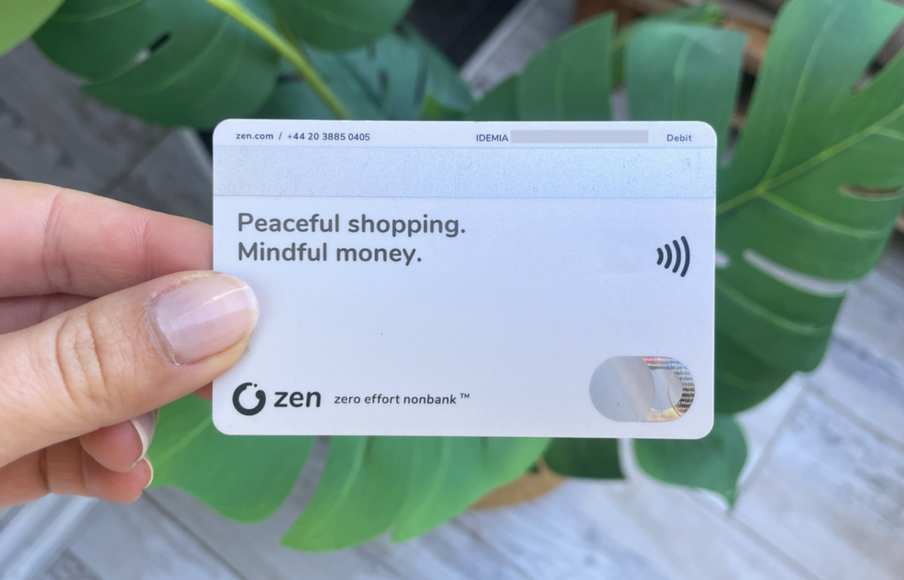 Osoba trzyma białą kartę płatniczą z logotypem "zen" nad napisem "Peaceful shopping. Mindful money." na tle roślin o zielonych liściach.