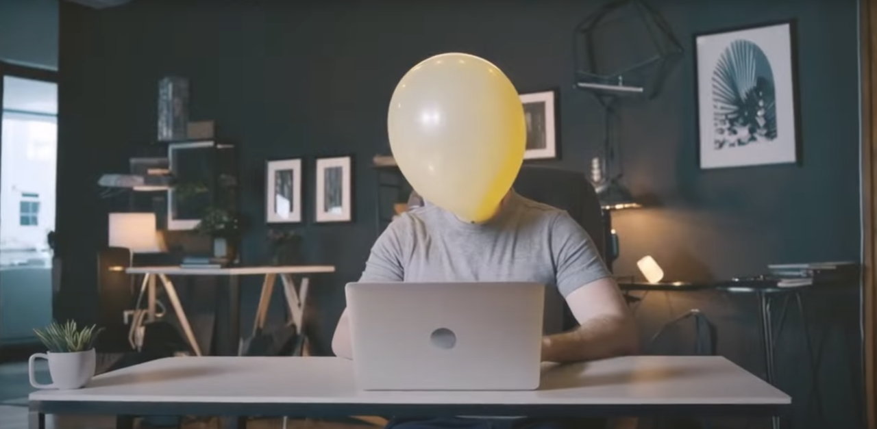 Osoba siedząca przy biurku z laptopem, z balonem na głowie zasłaniającym twarz, w stylowo urządzonym pokoju.