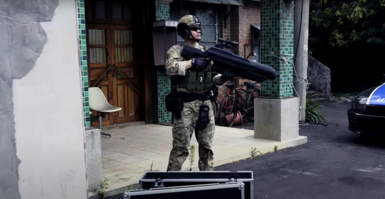 Zabójca dronów Skynet. Żołnierz w pełnym umundurowaniu trzymający duży granatnik, stojący przed budynkiem.