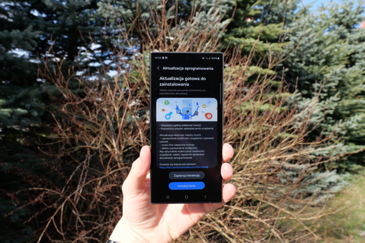 Mão segurando um smartphone com uma tela exibindo uma mensagem de atualização de software contra o fundo de árvores coníferas verdes naturais.