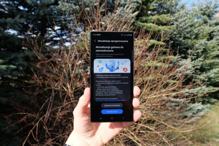 Ręka trzymająca smartfon z ekranem wyświetlającym komunikat o aktualizacji oprogramowania na tle naturalnej zieleni drzew iglastych.