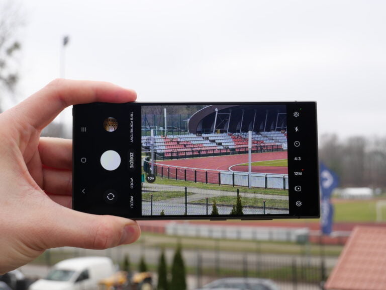 Ręka trzymająca smartfon, na którym widać zdjęcie stadionu lekkoatletycznego z bieżnią i trybunami.