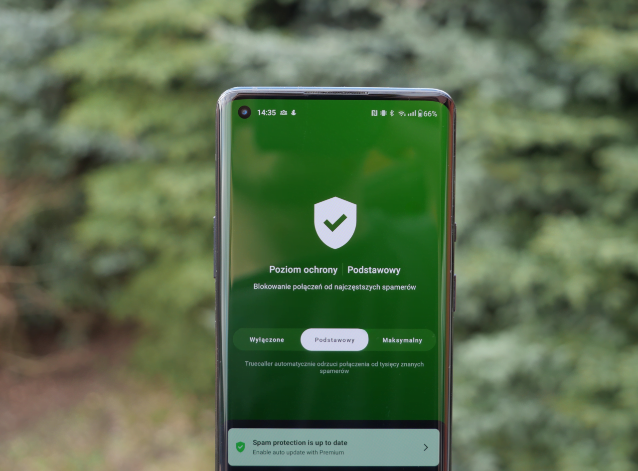 Smartfon trzymany przed tłem w postaci rozmazanych zieleni drzew, wyświetlając ekran ochrony przed spamem z zaznaczoną opcją "Podstawowy".